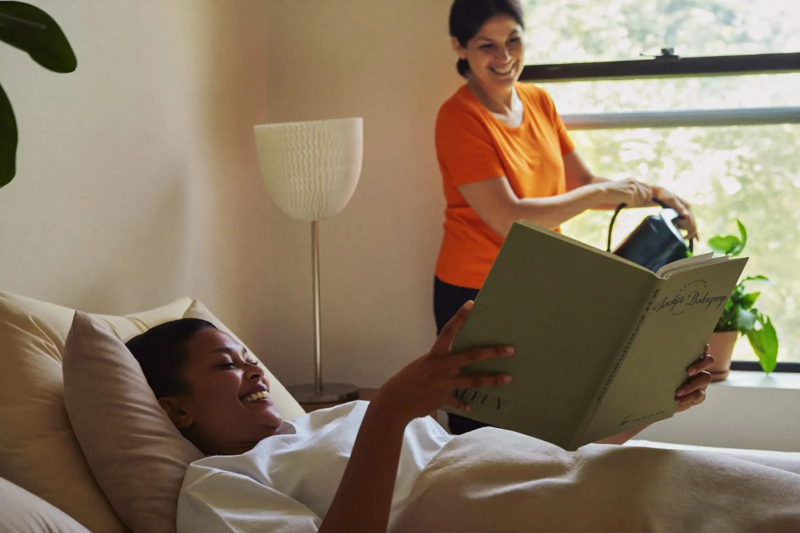 Junge Frau erholt sich zu Hause im Bett mit einem Buch, die Helferin gießt am Fenster eine Pflanze